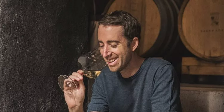 Wijnmaker Anton Baron Longo ruikt glimlachend aan een glas witte wijn in de wijnkelder van Baron Longo, met eikenhouten vaten op de achtergrond