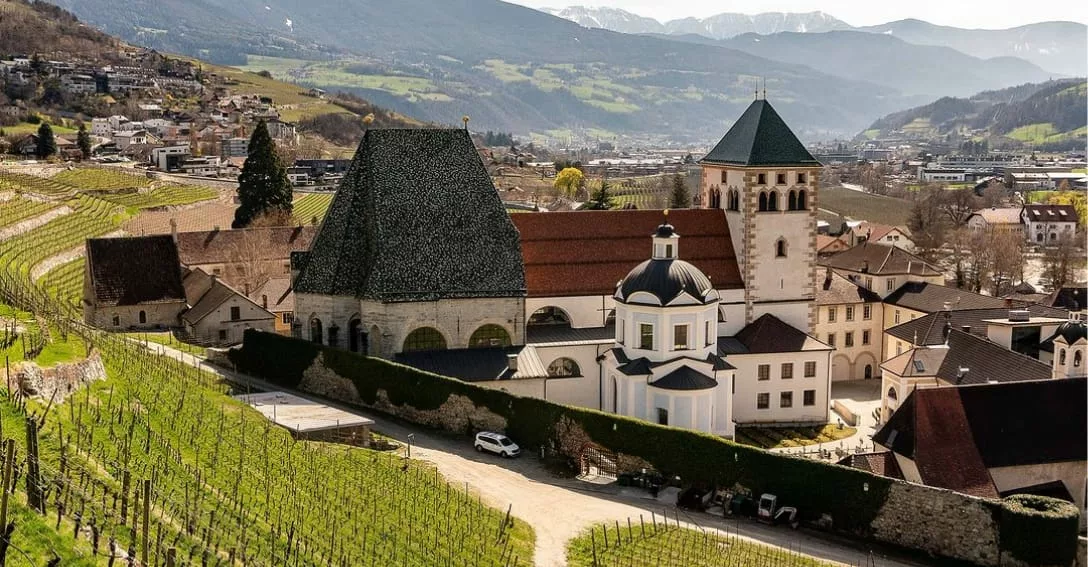 Historische Abdij van Novacella (Abbazia di Novacella) in Zuid-Tirol met stenen gebouwen, een markante toren met groen dak en een kerk met donkere koepel. Het complex ligt te midden van uitgestrekte wijngaarden in een bergvallei met besneeuwde toppen op de achtergrond.