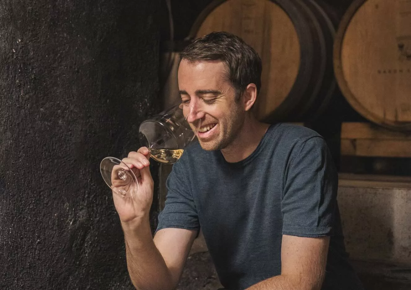 Wijnmaker Anton Baron Longo ruikt glimlachend aan een glas witte wijn in de wijnkelder van Baron Longo, met eikenhouten vaten op de achtergrond