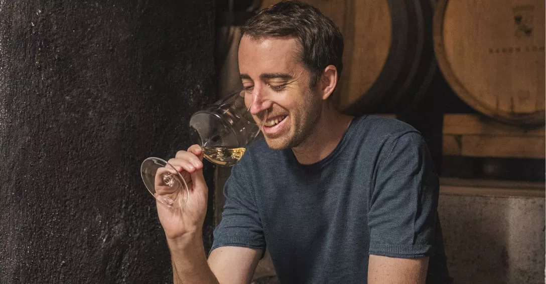 Wijnmaker Anton Baron Longo ruikt glimlachend aan een glas witte wijn in de wijnkelder van Baron Longo, met eikenhouten vaten op de achtergrond