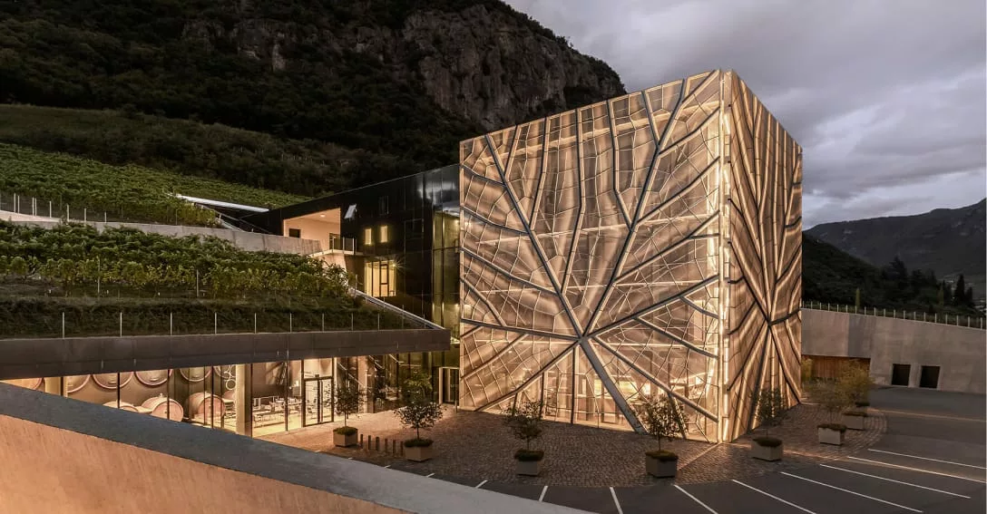 Moderne wijnmakerij Cantina Bolzano bij schemering. Het gebouw heeft een opvallend verlichte gevel met een gouden bladmotief en grote glazen gevels, gelegen aan de rand van wijngaarden met bergen op de achtergrond.
