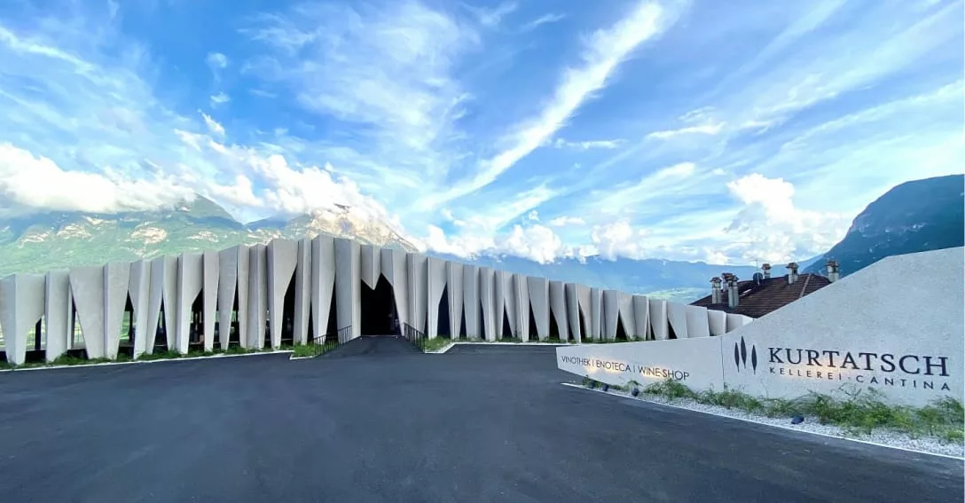 Moderne architectuur van Cantina Kurtatsch met kenmerkende betonnen verticale lamellen. Op de voorgrond staat het logo van de wijnmakerij, met bergen en een blauwe wolkenlucht op de achtergrond.