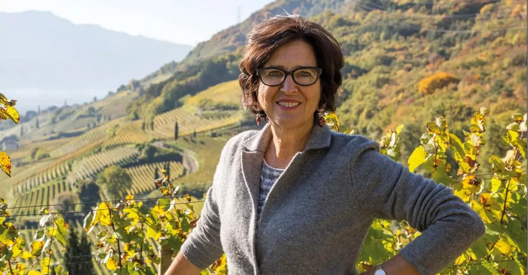 Portret van wijnmaakster Elena Walch in de herfstige wijngaarden van Zuid-Tirol. Ze staat met handen in de zij te glimlachen, omringd door kleurrijke herfstbladeren en heuvels met wijngaarden.